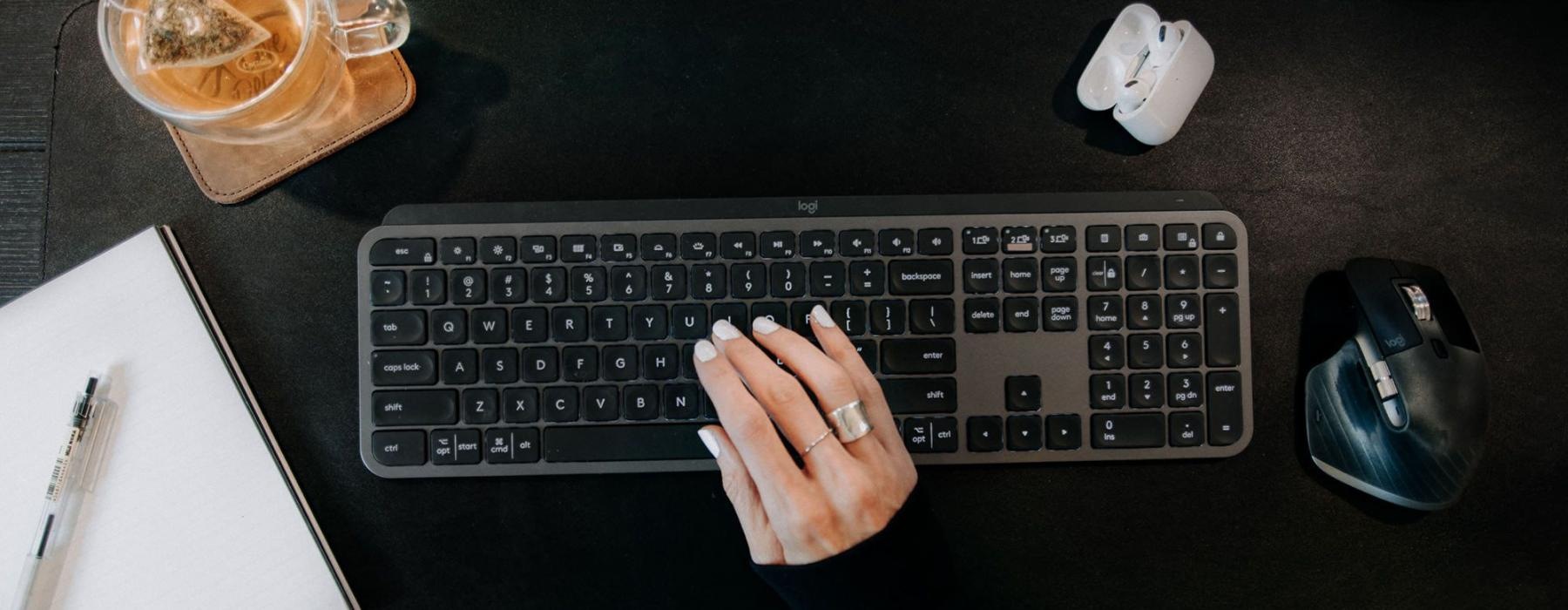 woman's hand on a keyboard surrounded by office items and a cup of tea on a coaster